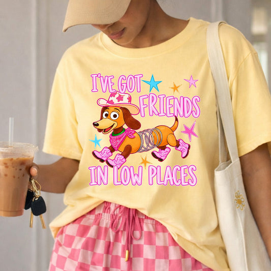High-quality DTF transfer on a bright blue t-shirt featuring a cartoon slinky dog wearing a pink cowprint cowboy hat, a pink bandana, and four pink cowboy boots. The text reads "I've Got Friends In Low Places" in bright retro pink font with colorful stars in the background. Ready to press heat transfer graphic.