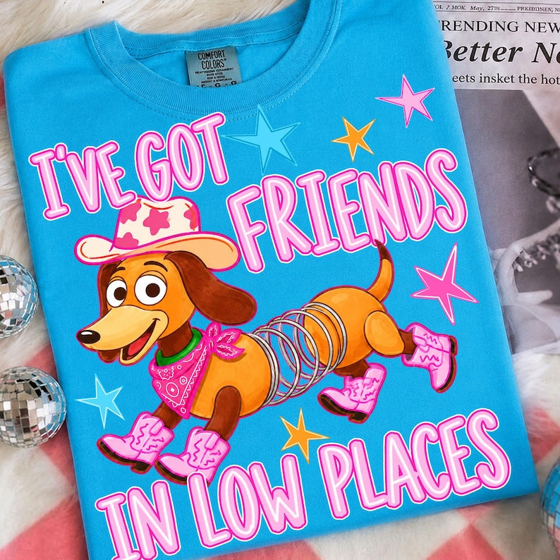 High-quality DTF transfer on a bright blue t-shirt featuring a cartoon slinky dog wearing a pink cowprint cowboy hat, a pink bandana, and four pink cowboy boots. The text reads "I've Got Friends In Low Places" in bright retro pink font with colorful stars in the background. Ready to press heat transfer graphic.
